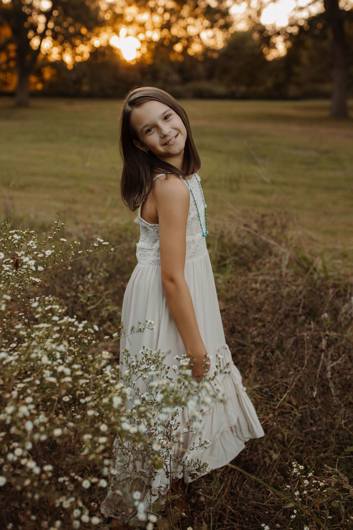 A person in a flowing white dress stands among wildflowers during golden hour sunset in a grassy field.