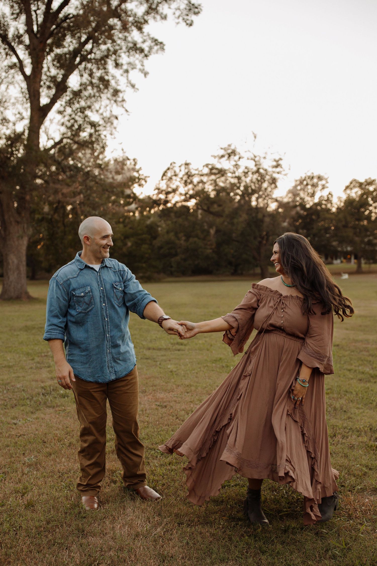 A couple in denim and brown attire dance playfully together in a grassy field at sunset.