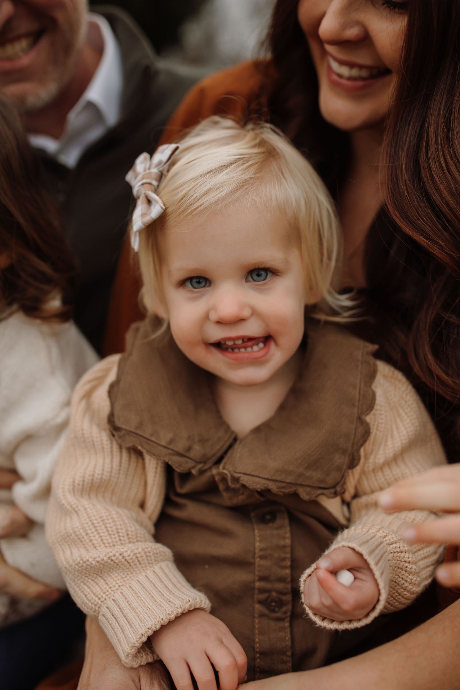 A toddler wearing a brown dress and beige sweater smiles while being held by family members in a warm portrait setting.