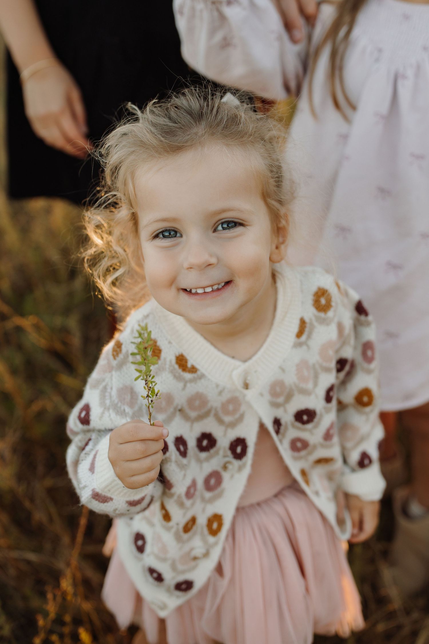 A young child in a floral blouse and pink skirt smiles while holding small flowers in a series of connected photos.