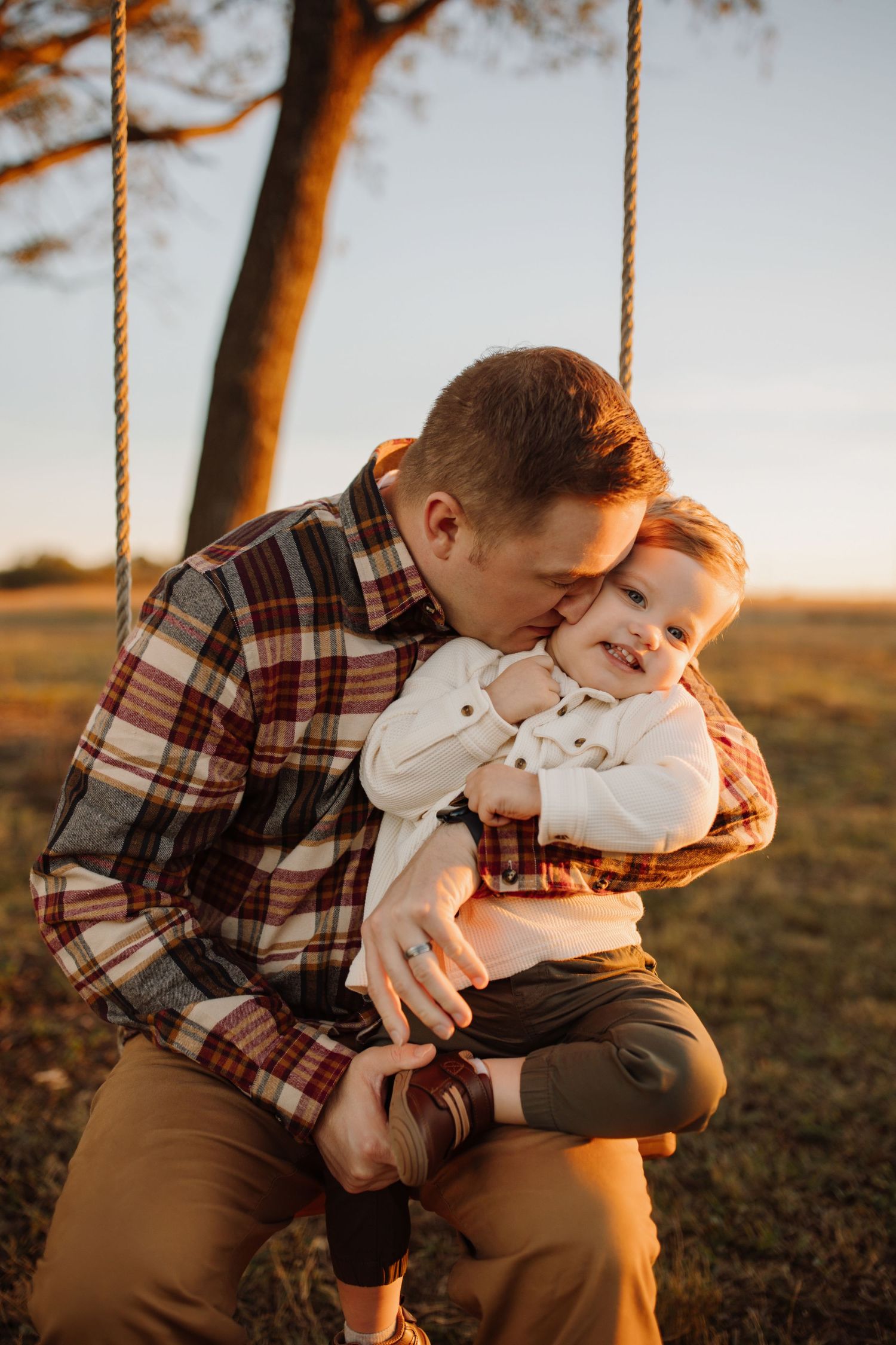 A tender moment captured at sunset as a parent and child share a swing in a plaid shirt and brown pants.