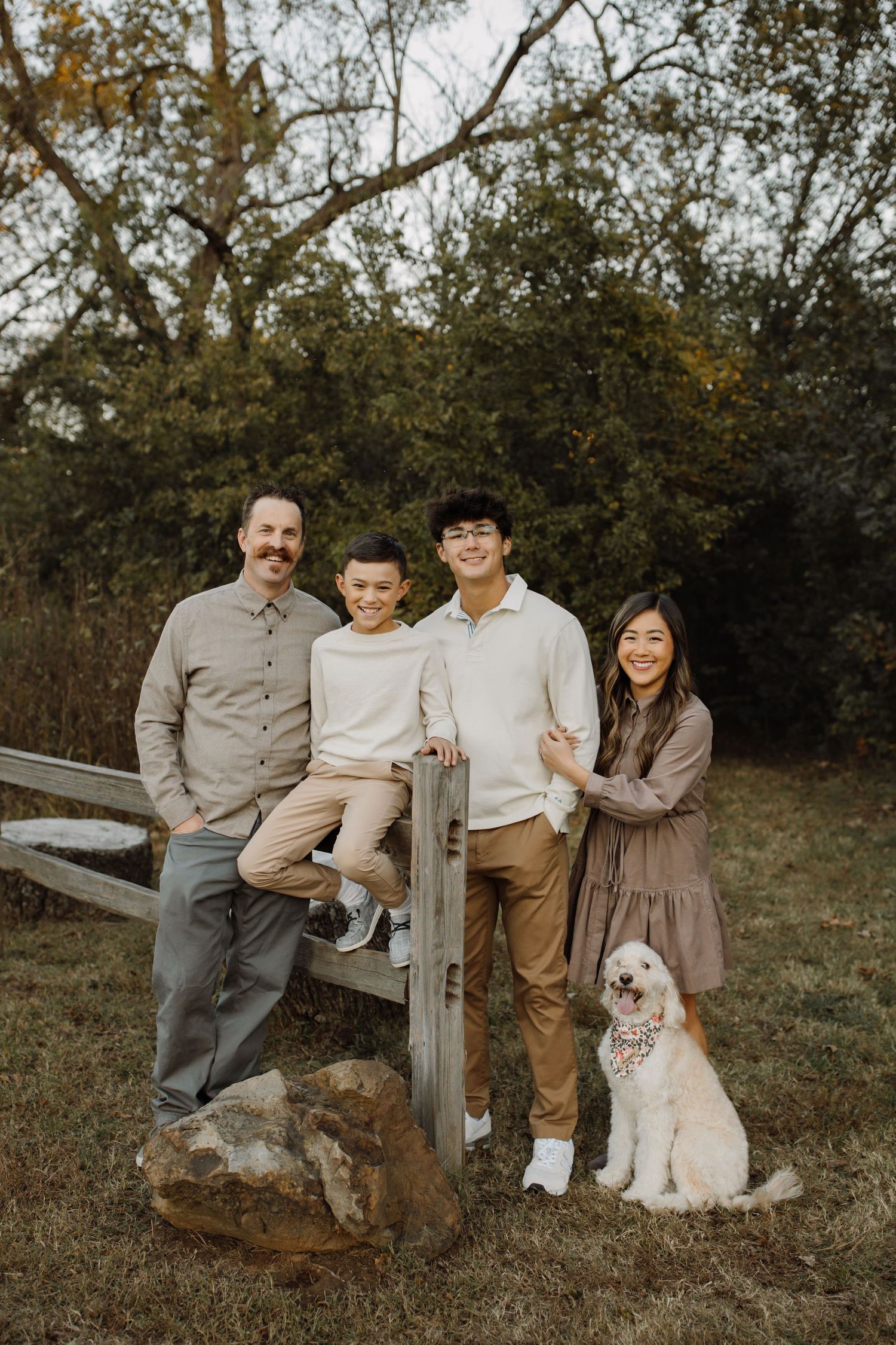 A family and their fluffy white dog pose together on wooden fence posts in a rustic outdoor autumn setting.