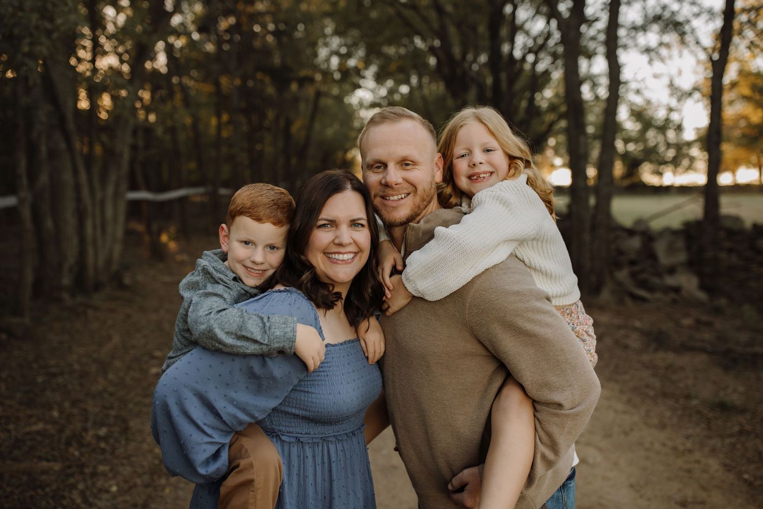 A family shares warm embraces and laughter during a golden hour photoshoot in a wooded setting.