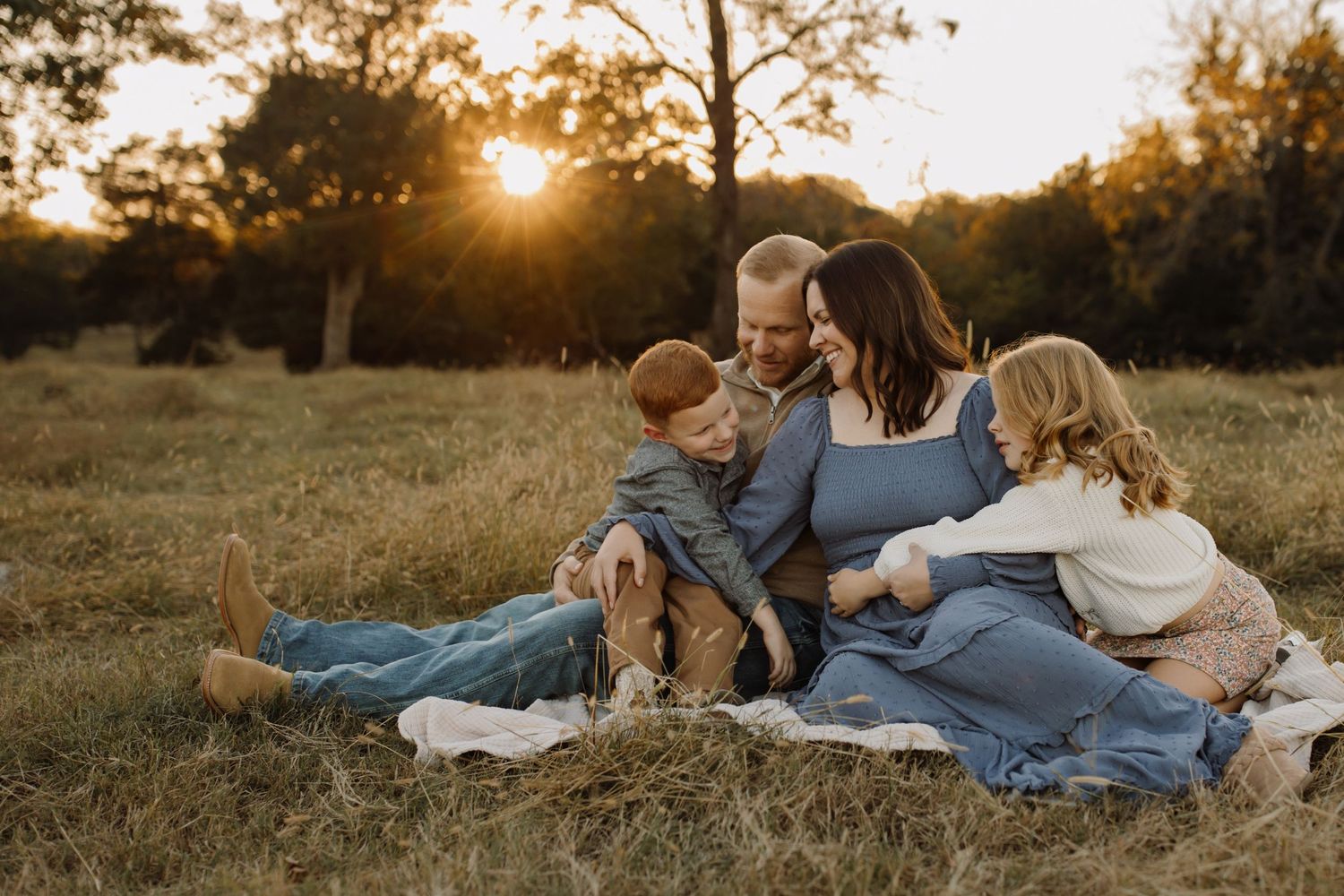 Family laying together on blanket in grassy field during golden sunset hour.