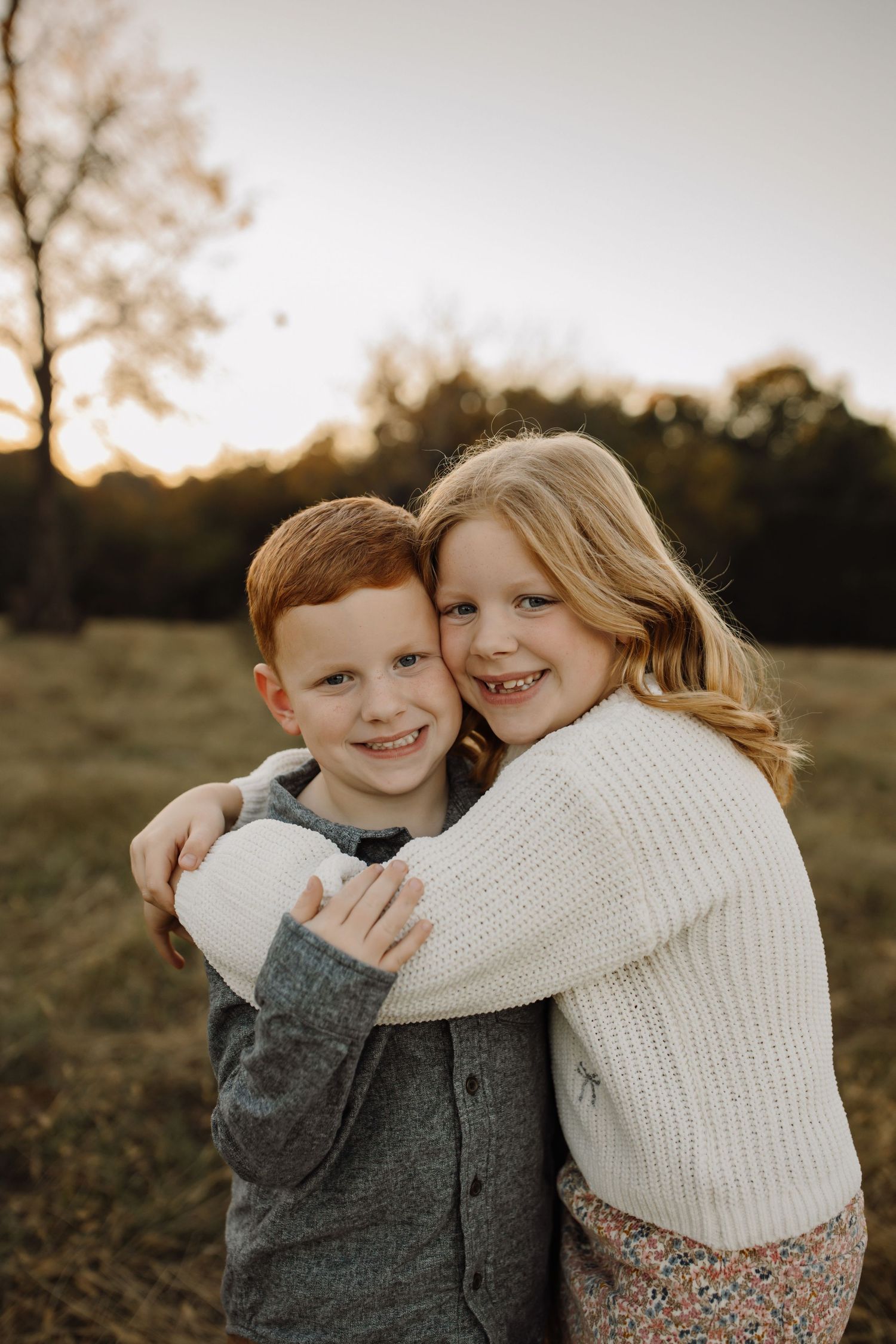 Two young children in warm sweaters embrace and smile while sitting together in a grassy field at sunset.