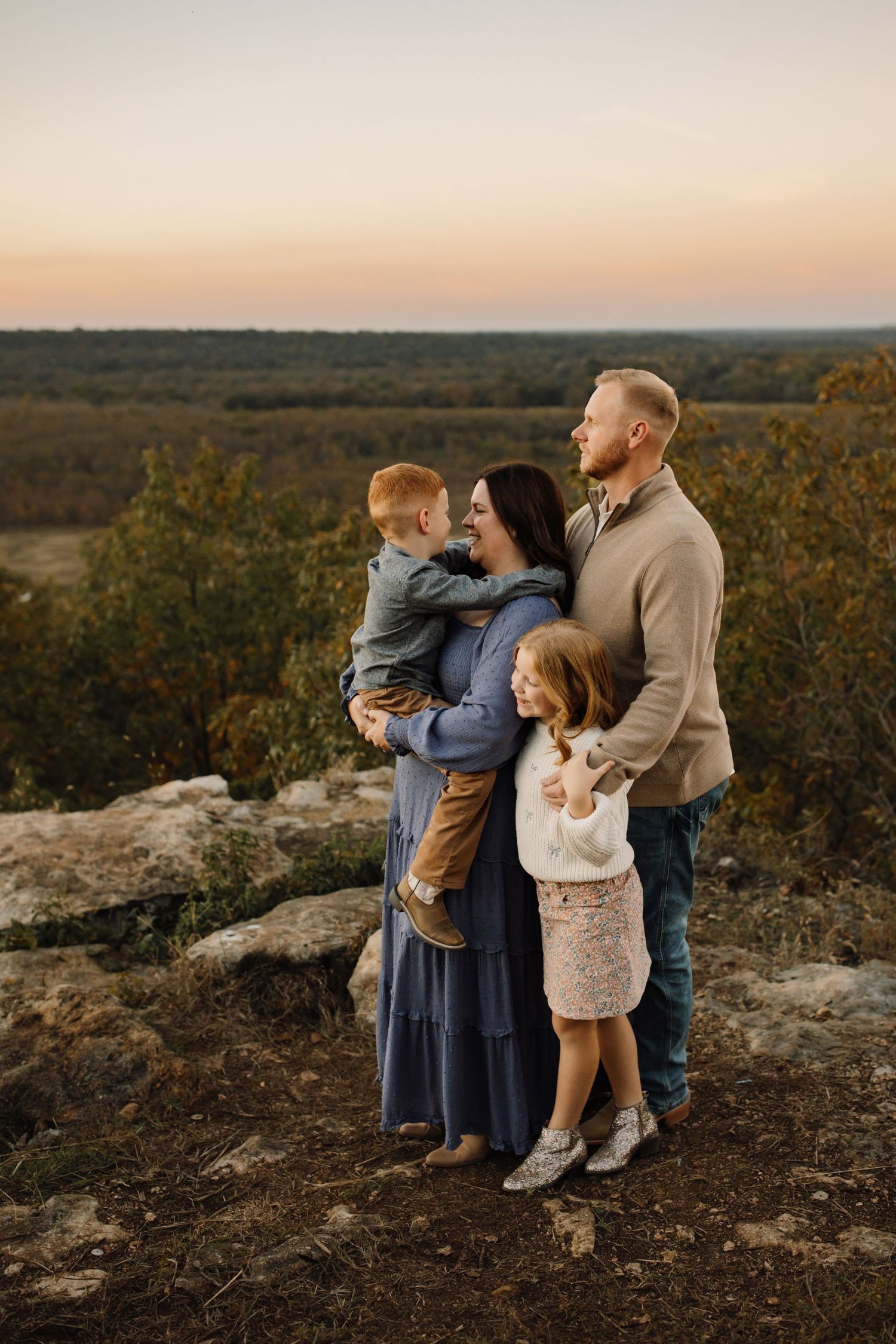 A family shares tender moments together during sunset on a rocky hilltop overlooking a scenic autumn landscape.