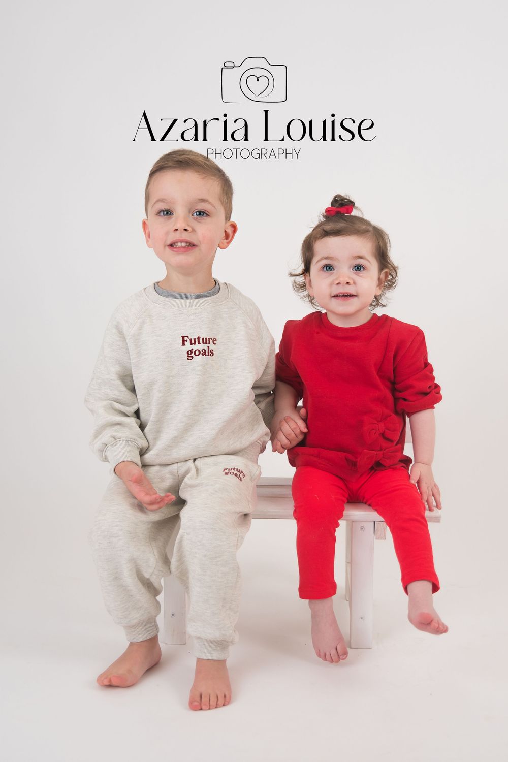 Professional studio portrait of two young children in matching loungewear outfits on a white bench against white background.