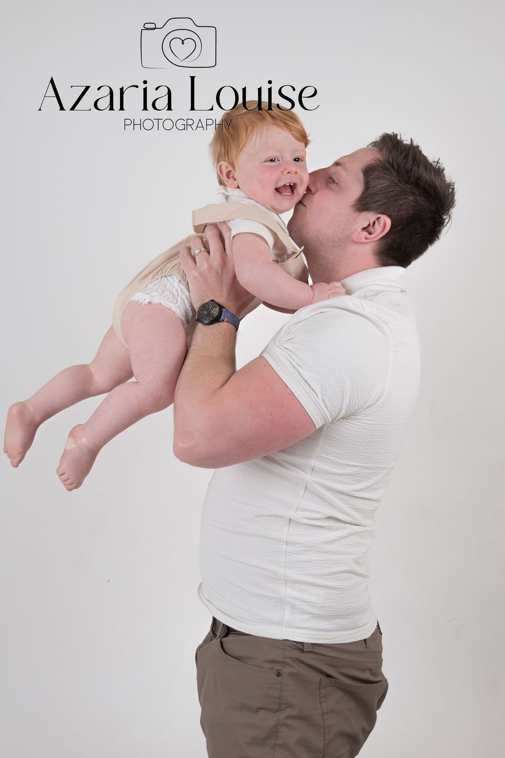 Professional studio photography of parent in white shirt lifting joyful infant in diaper against light backdrop.