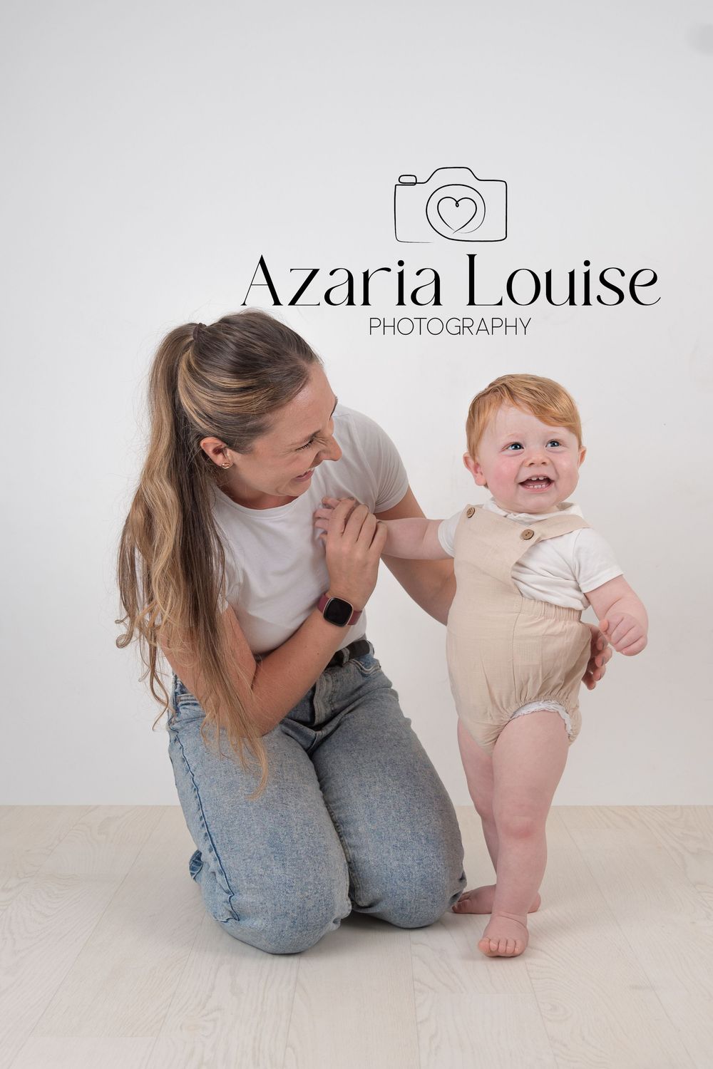 A photography studio session showing a mother in jeans kneeling with her baby in a beige romper against a white backdrop.