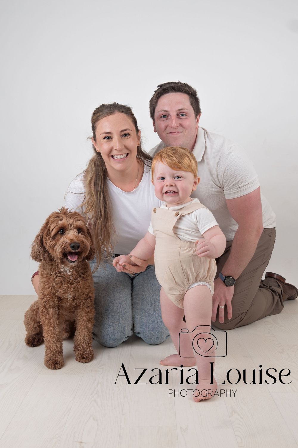 Family portrait with brown goldendoodle dog and baby in white outfit against light studio background.