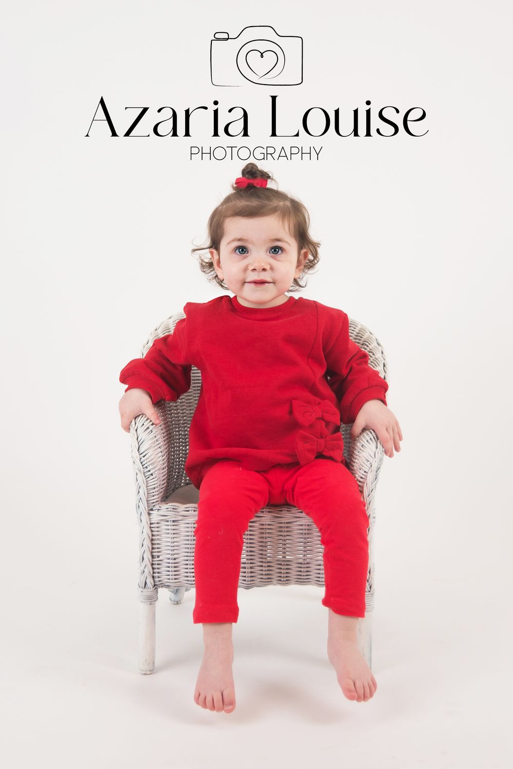 Studio photo showing a small child in bright red outfit sitting on a white wicker chair against white background.
