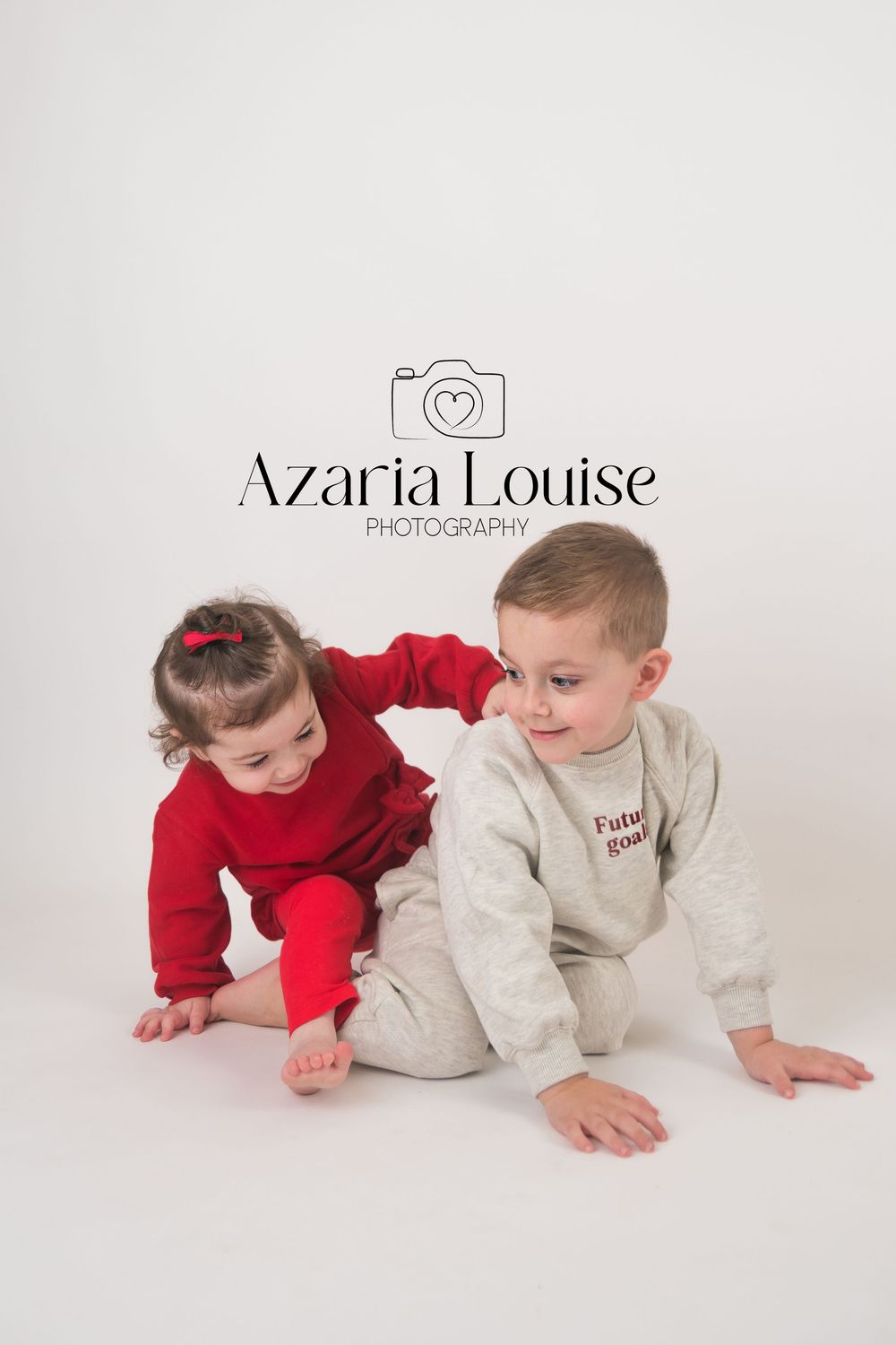 Two young children in red and beige sweaters kneel on a white backdrop during a professional photo studio session.