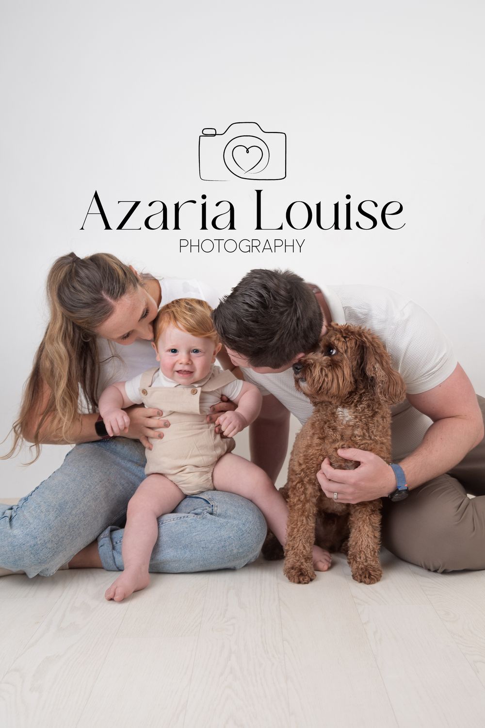 A heartwarming family photo session with parents, baby, and their brown goldendoodle dog on a light background.