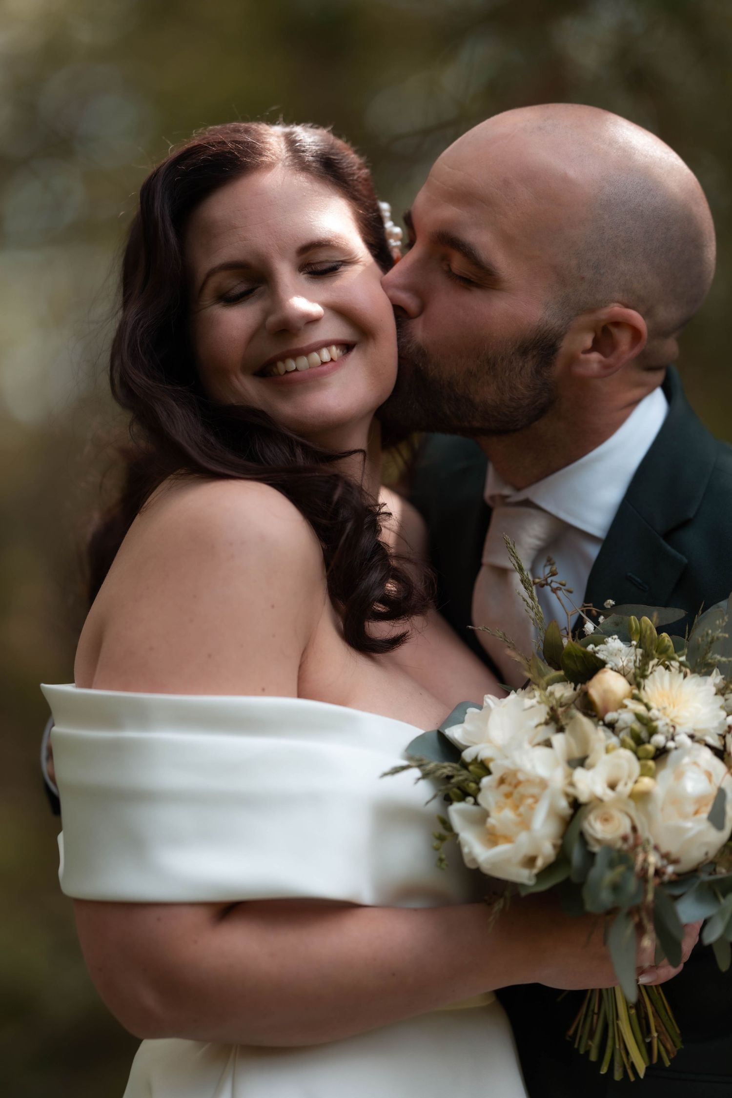 Smiling bride in off-shoulder white gown holds roses as bald groom kisses her cheek outdoors.