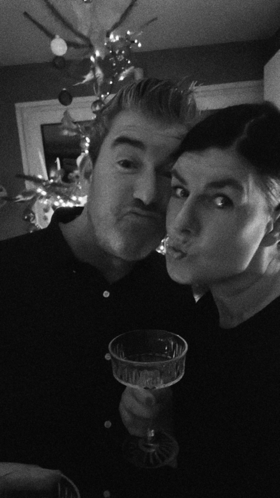 A black and white selfie of two people making faces while holding a cocktail glass with a Christmas tree in background.