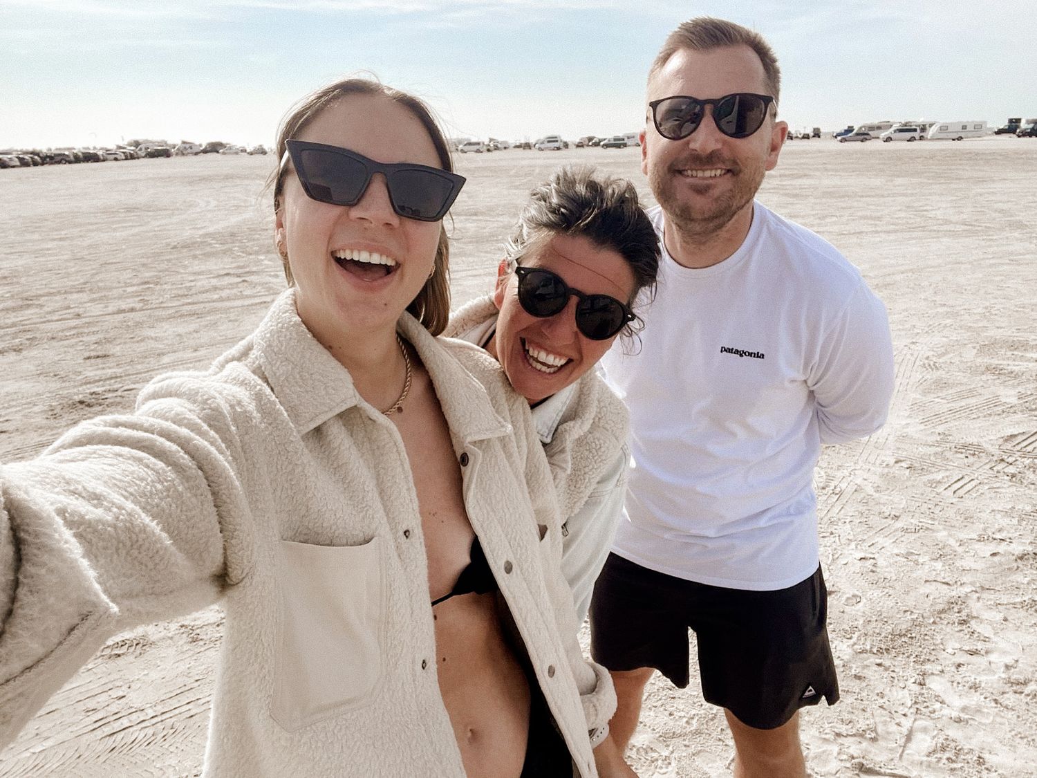 Three friends wearing sunglasses take a selfie together on a sunny desert landscape.