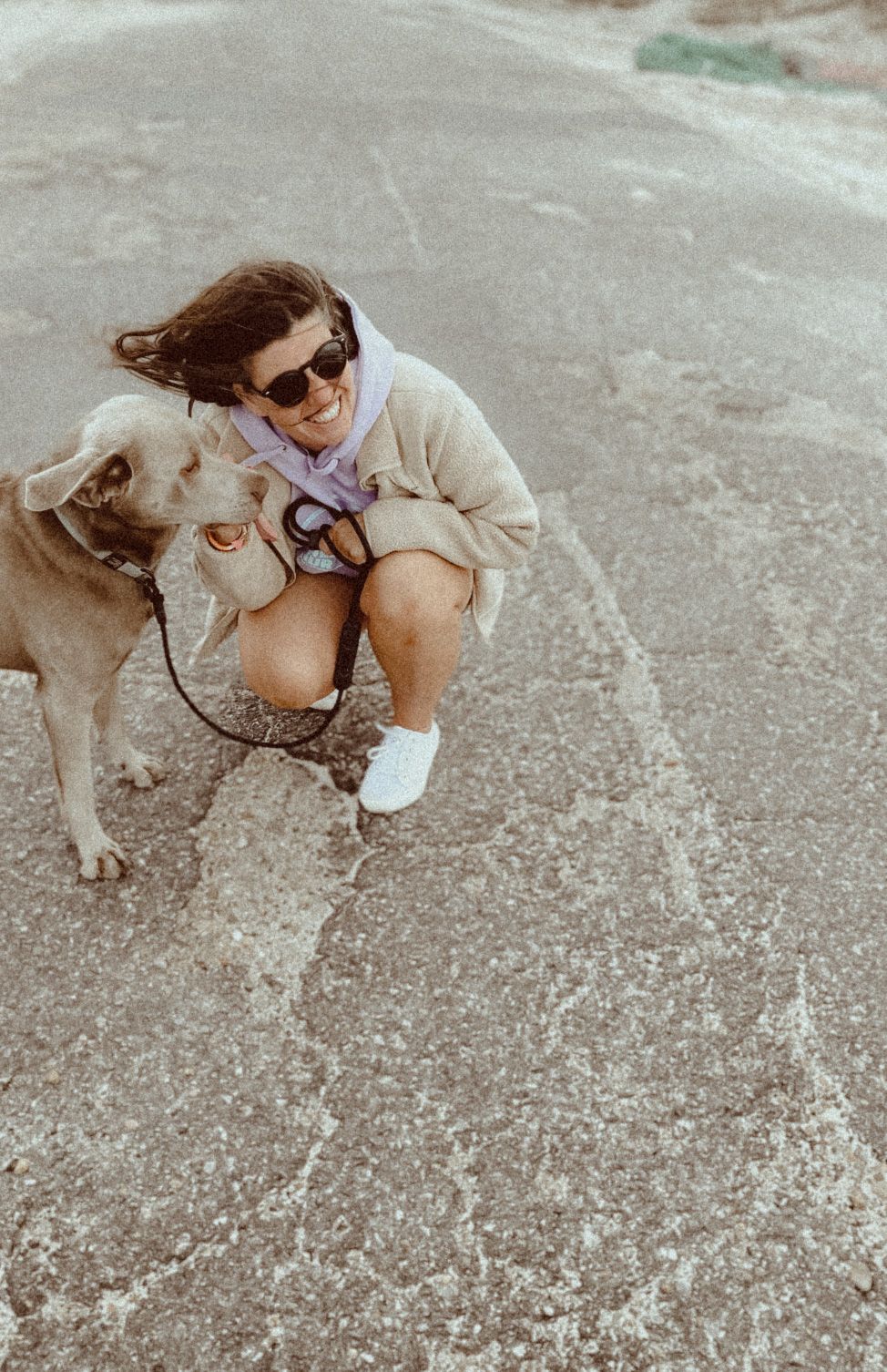 A person in a cream sweater and sunglasses crouches next to their golden-colored dog on a concrete surface.