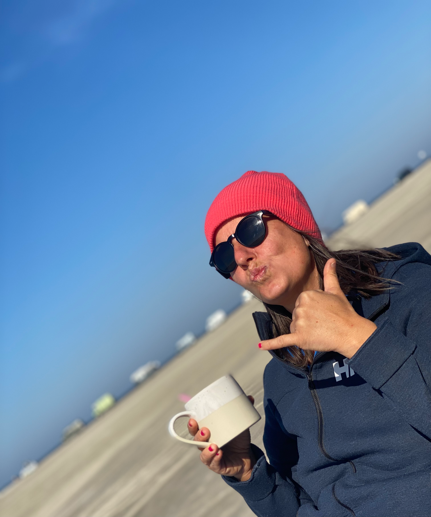 Person in red beanie and sunglasses holds a coffee cup while giving thumbs up against blue sky background.