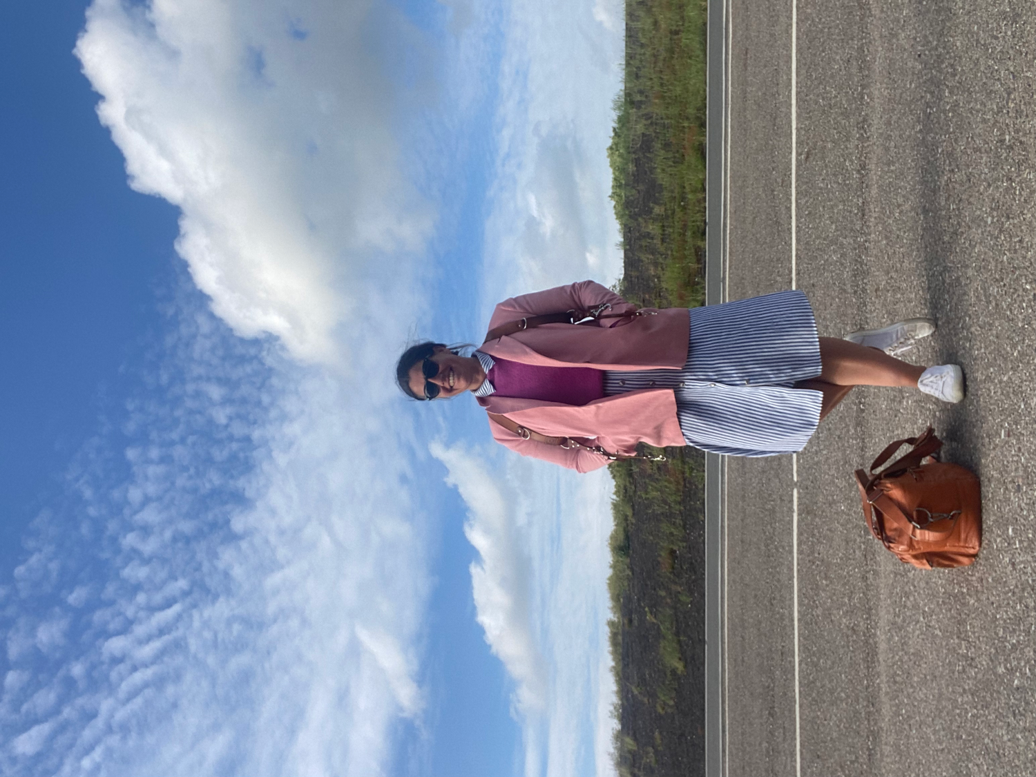 Someone wearing a pink jacket and white shorts stands on a paved path under a bright blue sky with puffy clouds.