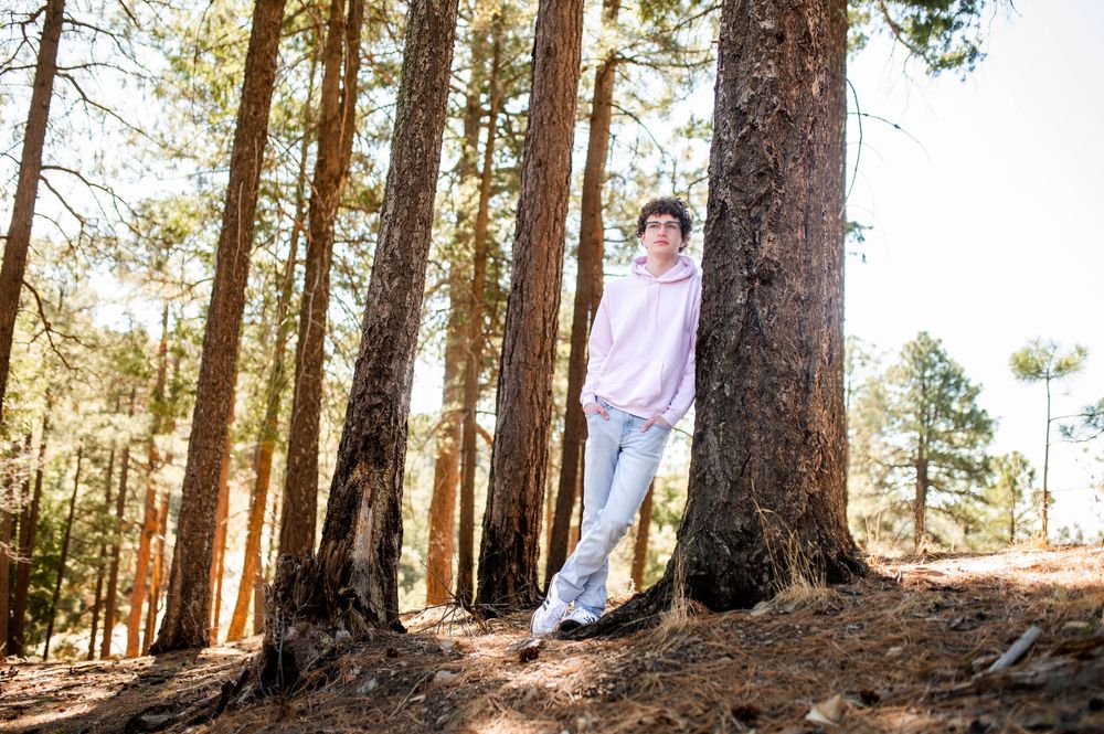 A young man in a pink hoodie and gray joggers leans casually against a large pine tree in a forest.