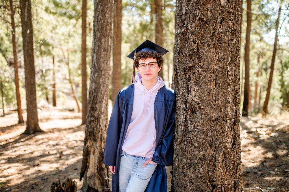 A high school graduate in cap and gown leans against a pine tree in a sunlit forest setting.