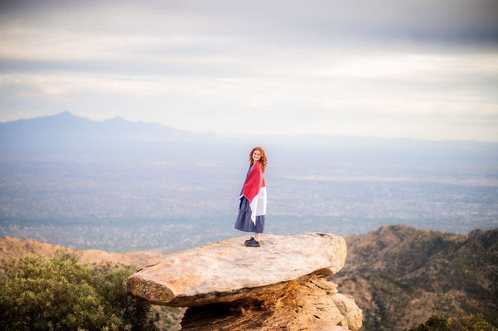 Woman in red top stands on rocky outcrop with vast mountain valley landscape behind her.