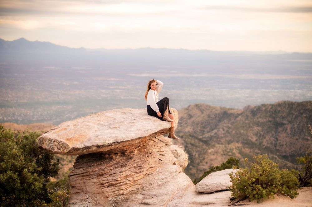Woman sitting on a dramatic rock ledge overlooking a vast valley and mountain landscape at sunset.