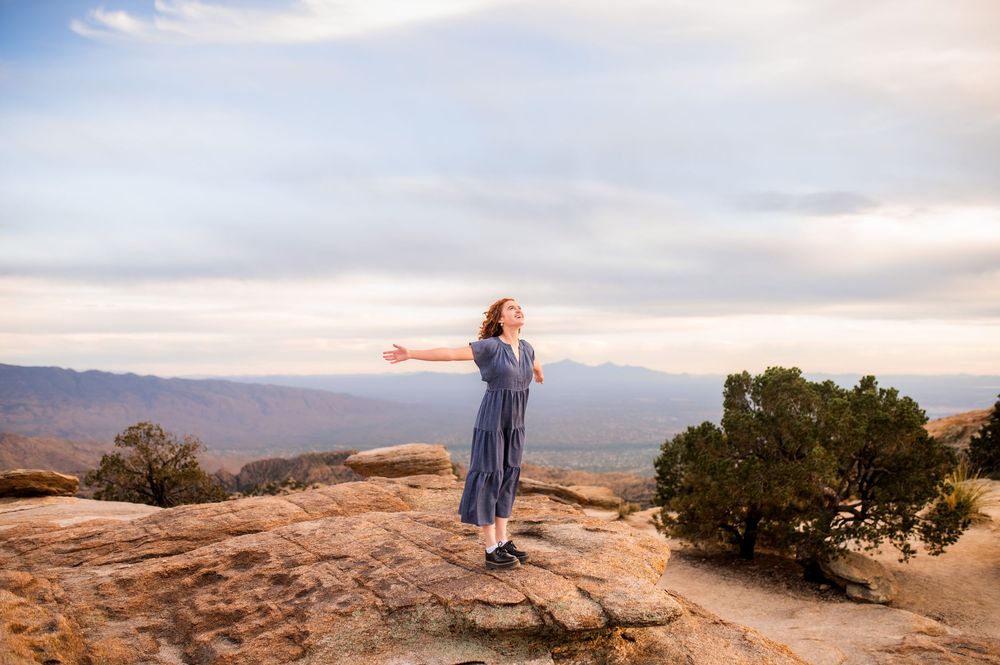 Woman in blue dress standing on red rock formation with arms outstretched, overlooking a vast desert valley.