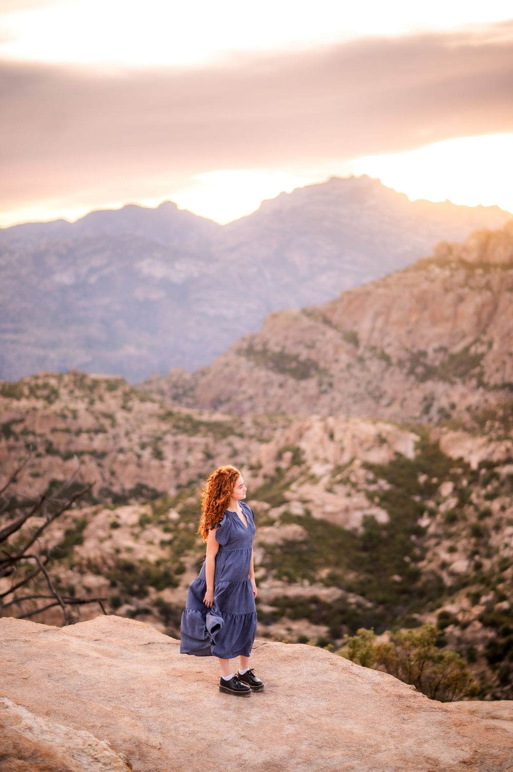 A red-haired woman in a blue dress stands on a rocky ledge overlooking a mountainous desert landscape at sunset.
