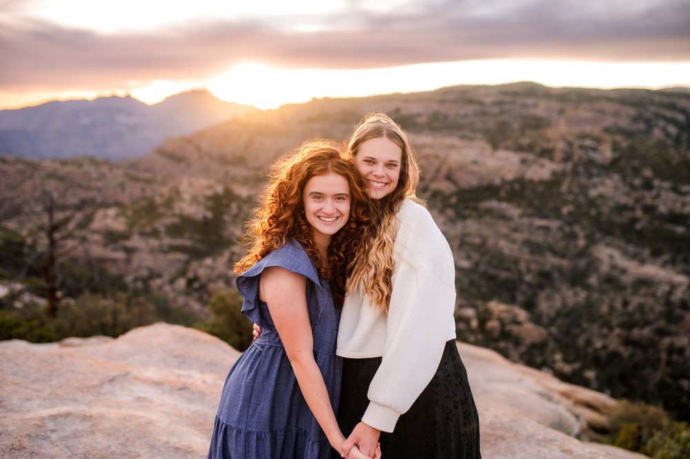 Two smiling young women holding hands on a rocky overlook with a stunning mountain sunset backdrop.