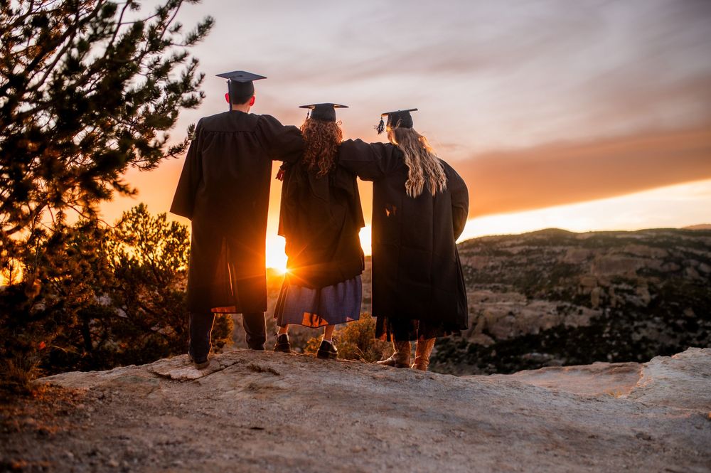 Three graduates in caps and gowns stand together on a rocky overlook, watching a stunning sunset over a scenic landscape.