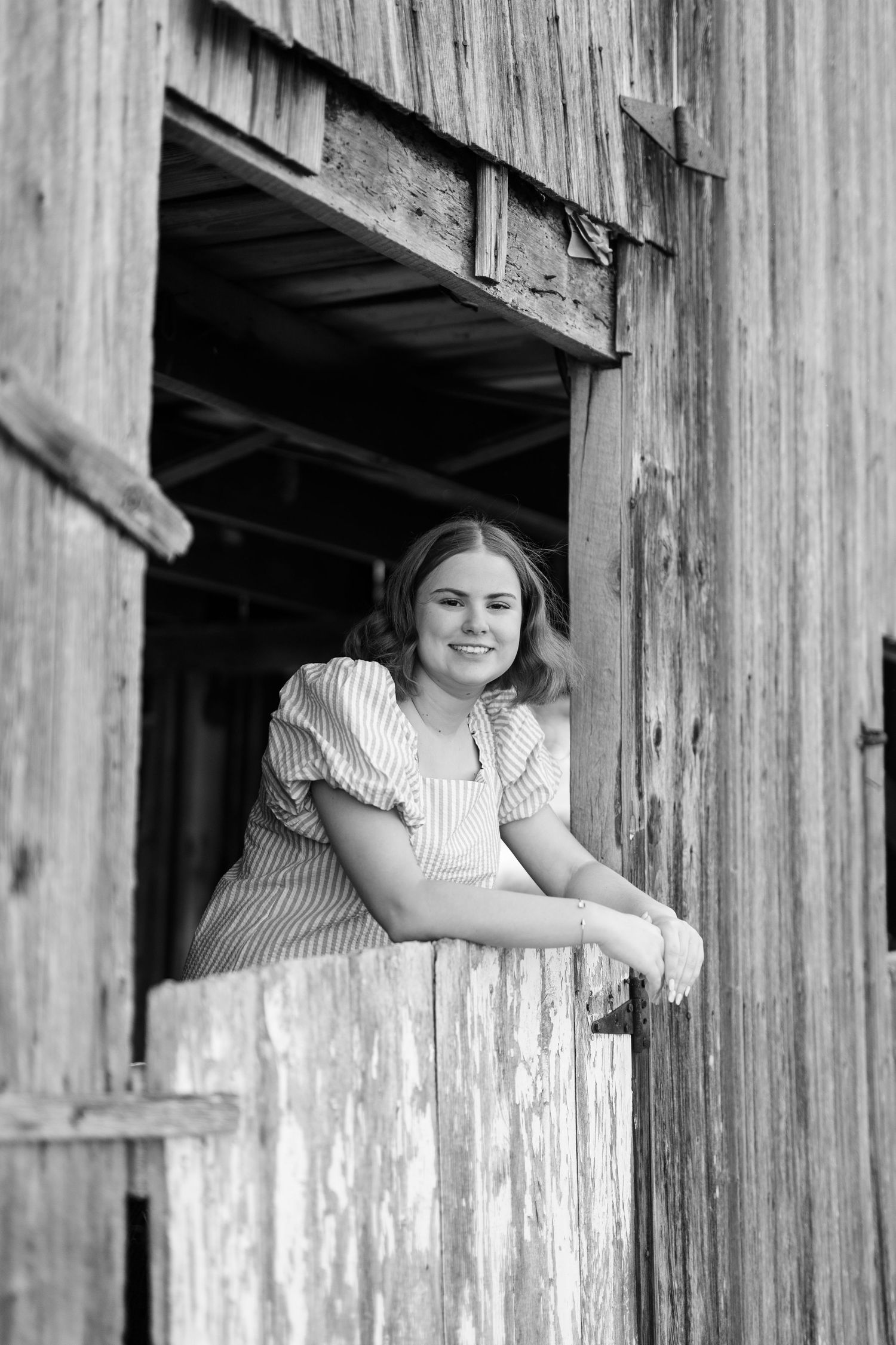Smiling young woman in white sweater leaning on fence post near rustic barn and silo.