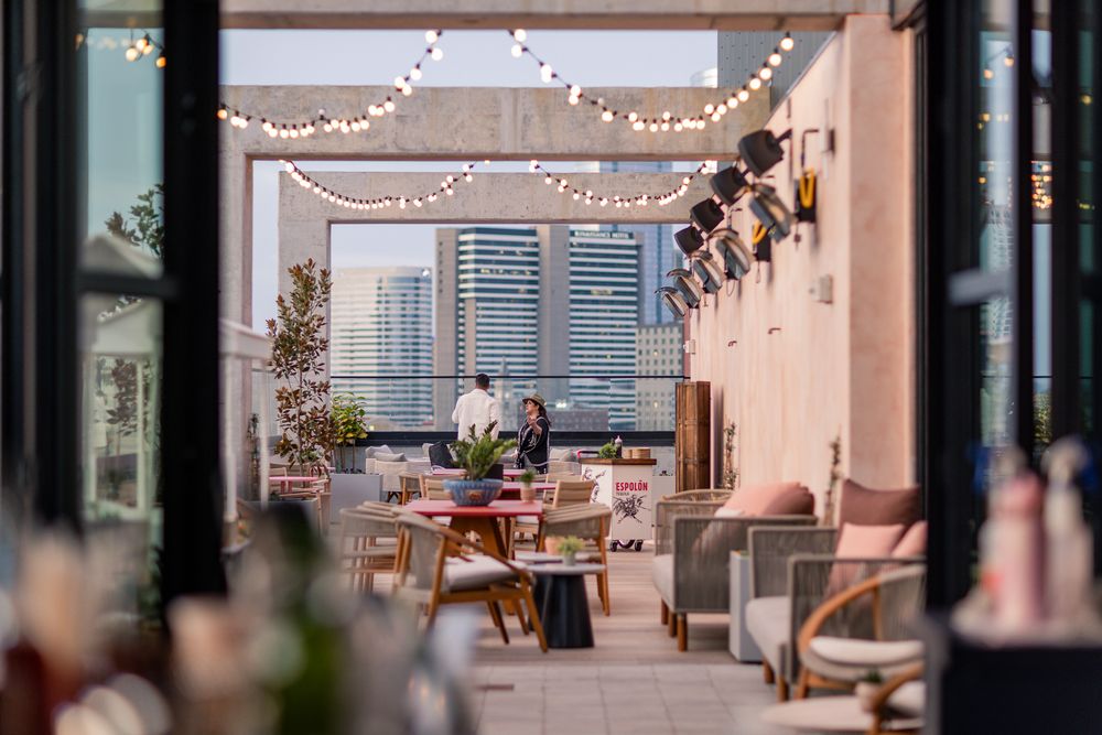 Rooftop restaurant patio with string lights, modern furniture, and city skyline views on a sunny day.