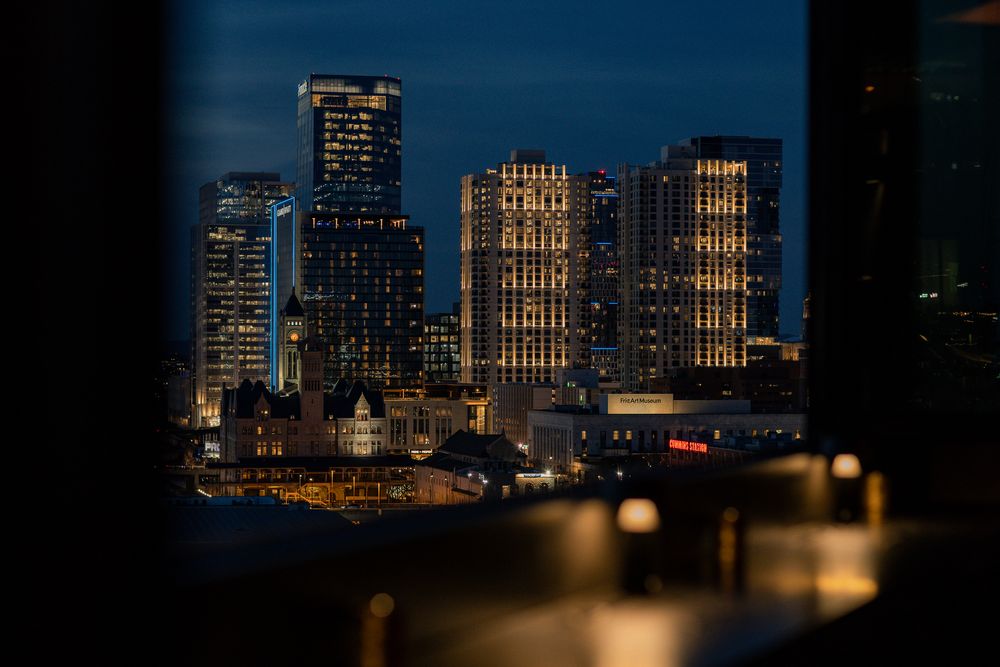 Night cityscape view through window frame showing illuminated skyscrapers and historic buildings against dark blue sky.
