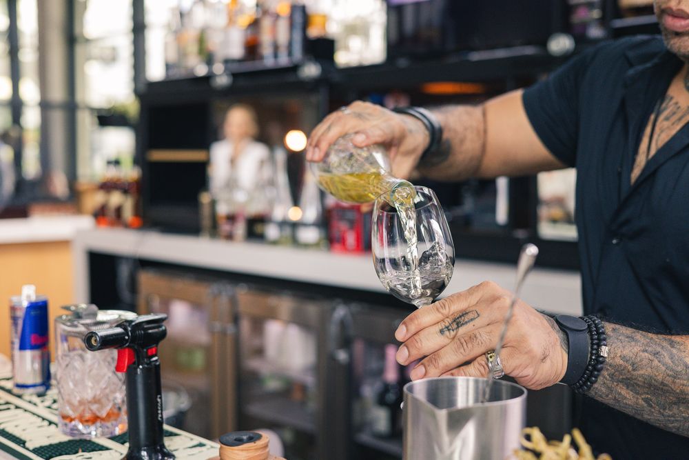 Tattooed bartender pouring a cocktail into a glass at a modern bar with bottles in the background.