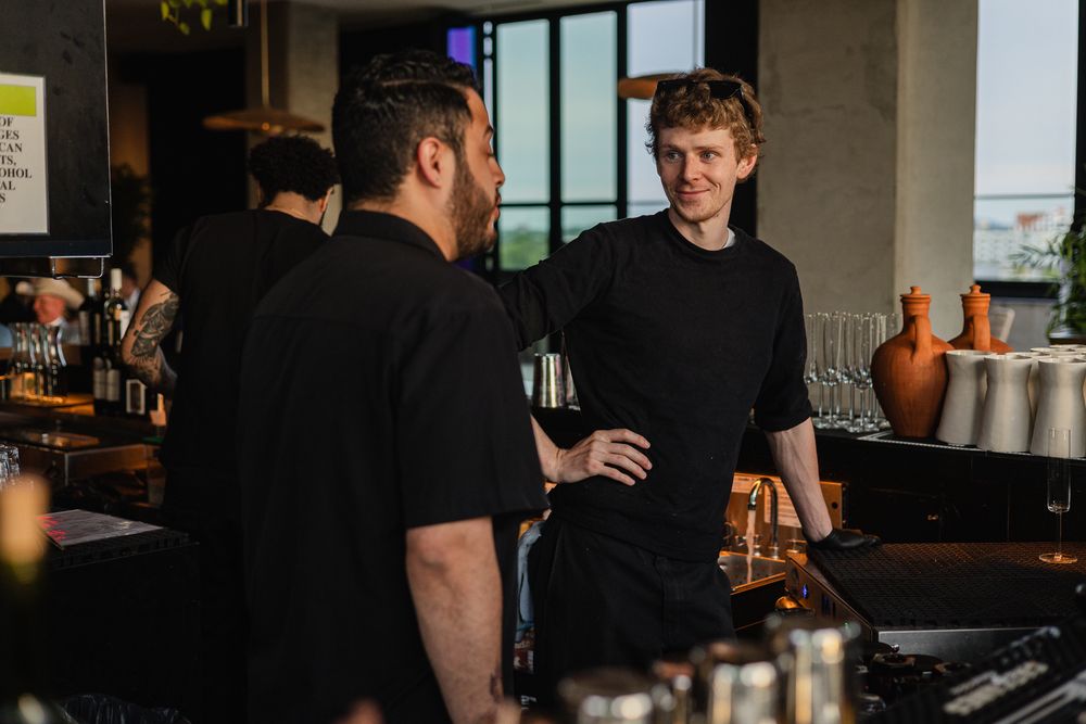 Two bartenders in black uniforms conversing at a modern bar counter in a stylish restaurant setting.