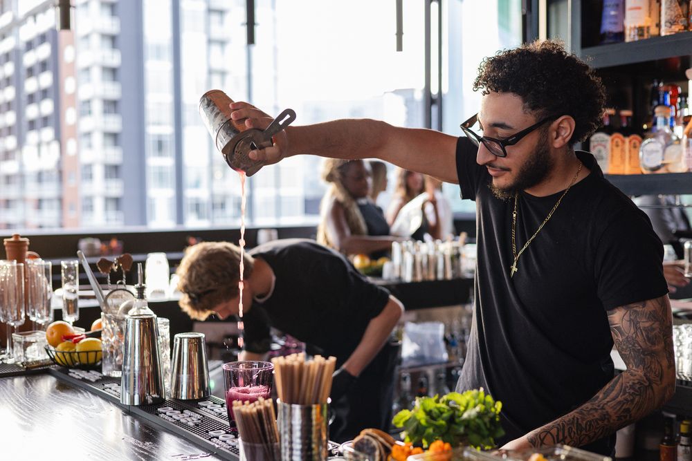Tattooed male bartender pouring cocktail shaker at a rooftop bar with city skyline in background.