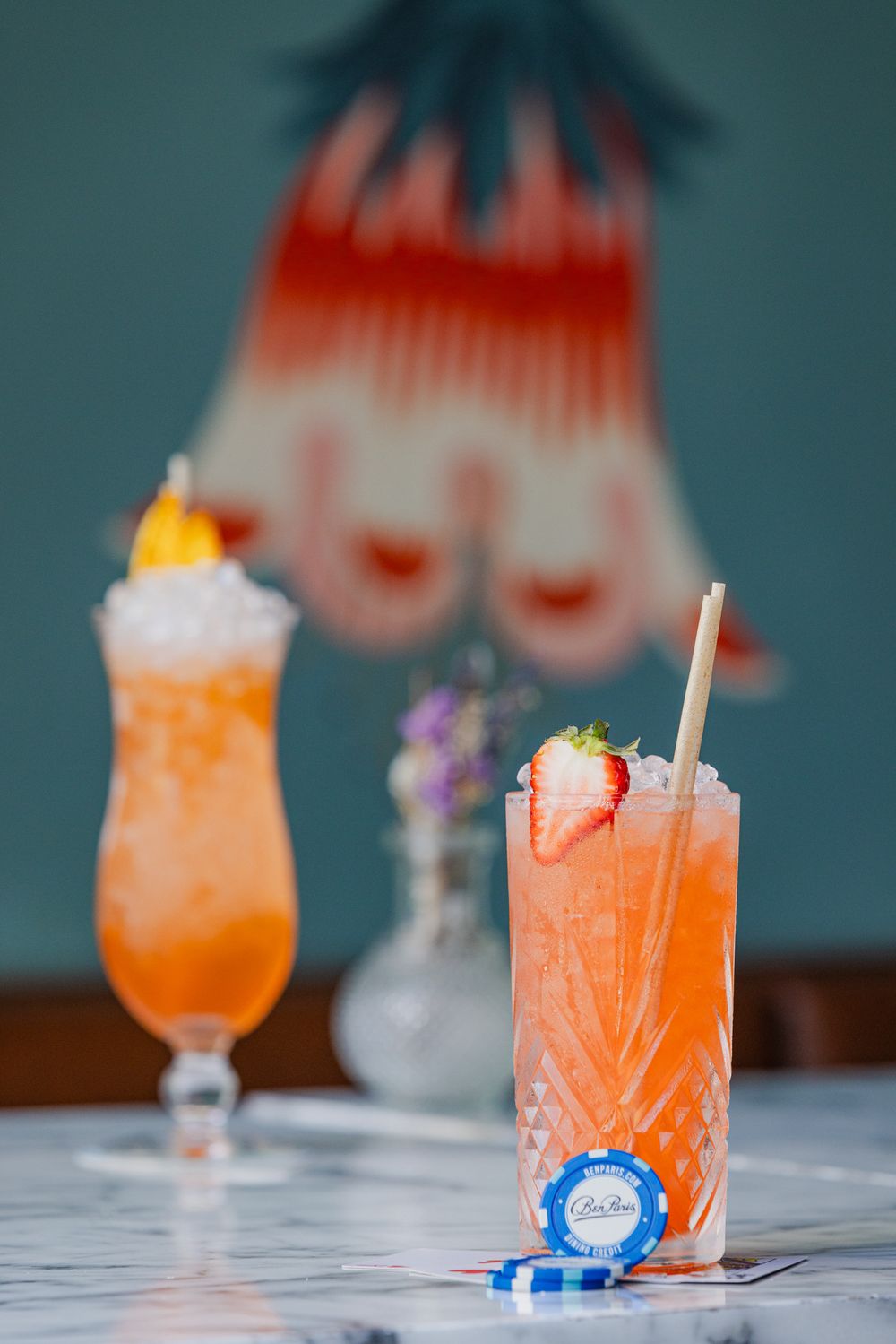 Two colorful tropical cocktails garnished with strawberries and whipped cream sit on a marble counter at a bar.