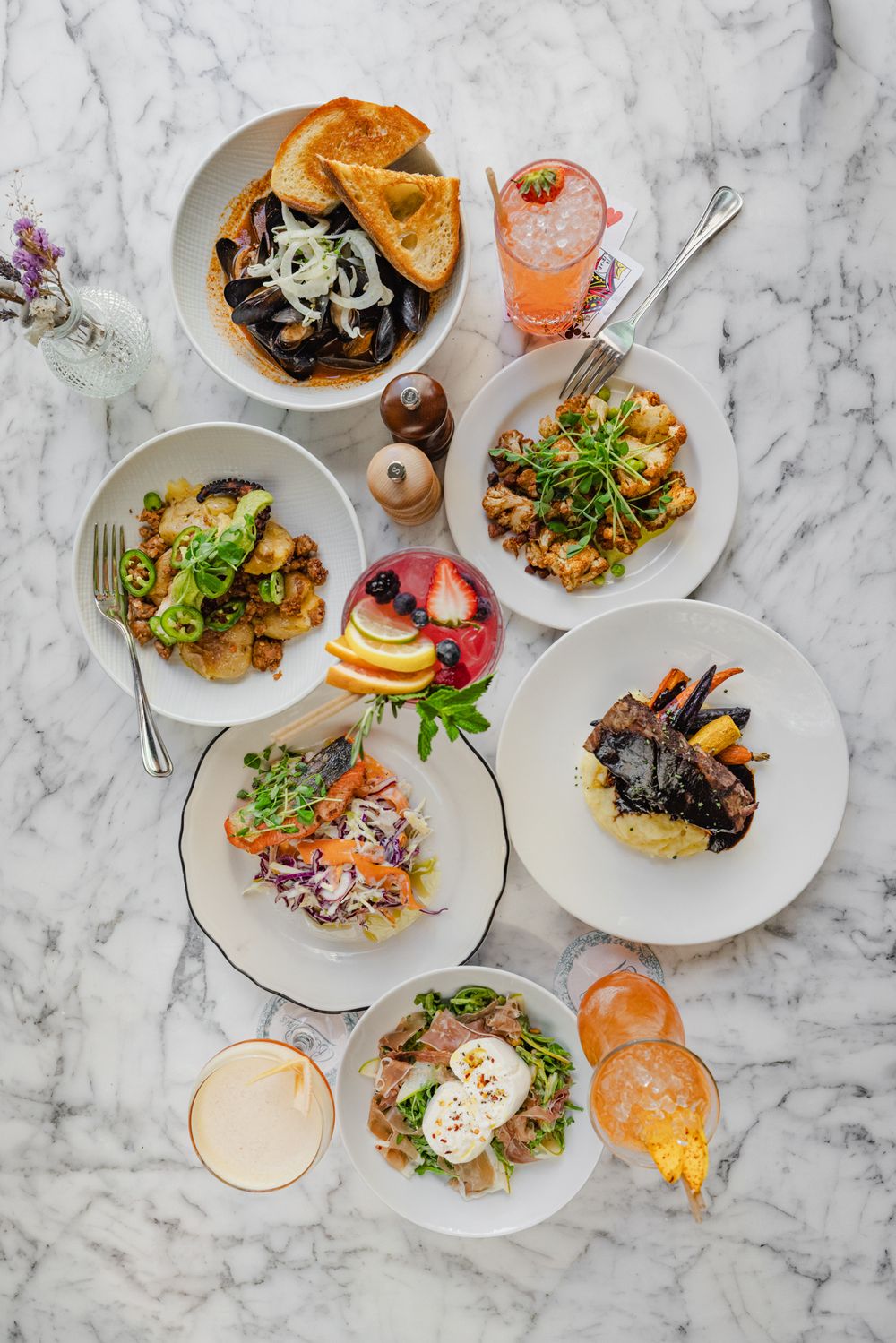 Aerial view of various brunch dishes including salads and sandwiches on white plates against a marble table surface.