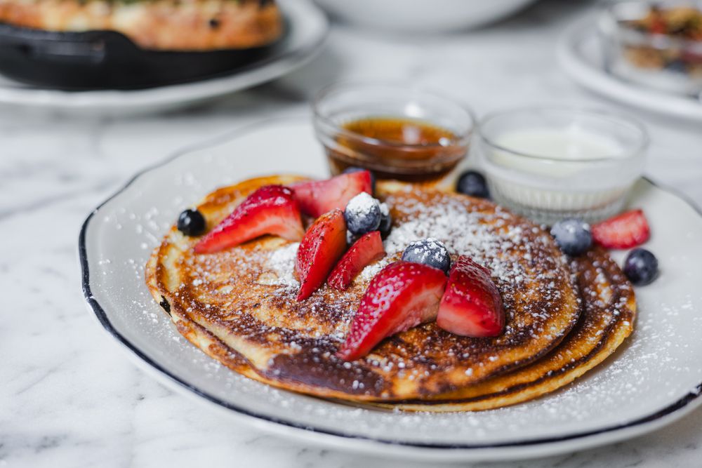 Stack of pancakes topped with fresh strawberries, powdered sugar, and served with maple syrup and cream.