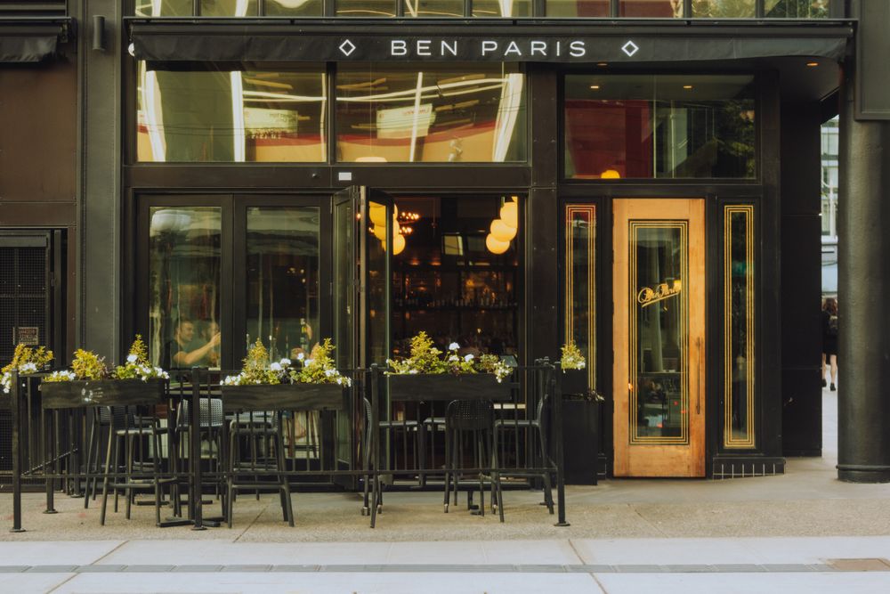 A wide view of modern mixed-use urban retail storefronts and sidewalk cafes with outdoor seating in the daytime.