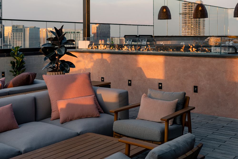 Rooftop lounge at sunset with gray sofas, wood chairs, pink cushions, pendant lights, and city skyline views.