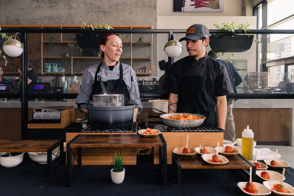 Two chefs in aprons prepare dishes at a rustic wooden kitchen counter in a modern restaurant setting.