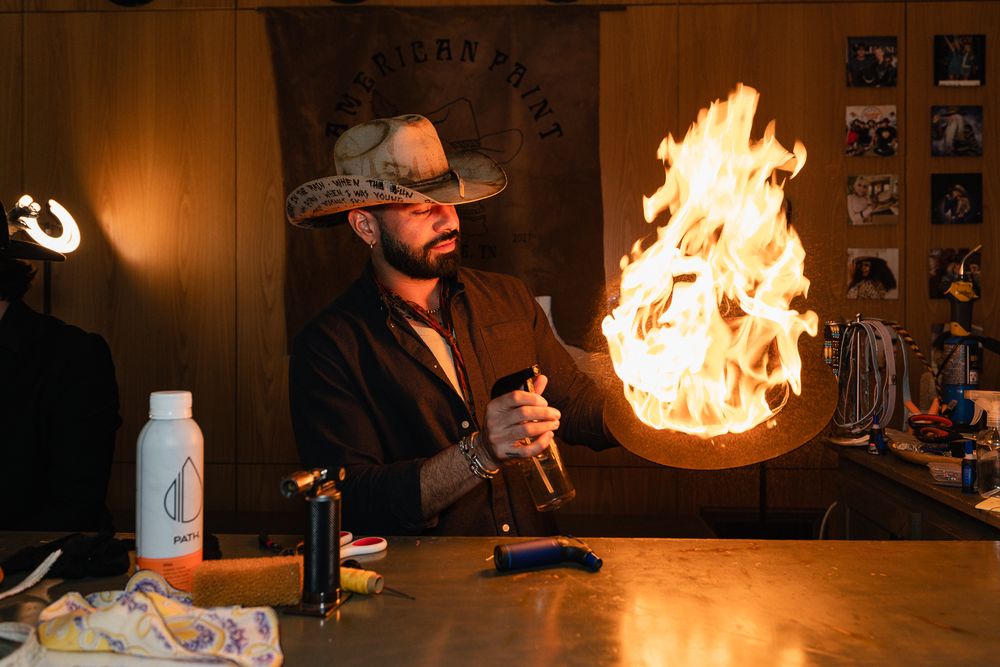 Man in cowboy hat performing fire trick with bottle at rustic bar counter indoors.
