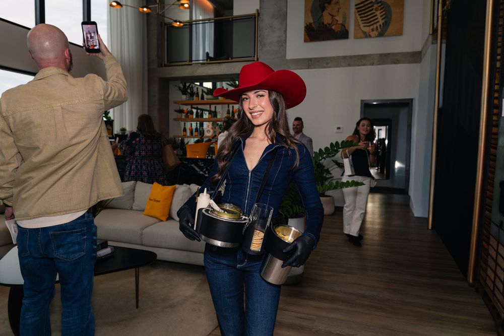 Woman in red cowboy hat and denim outfit smiling while holding champagne bottles at an indoor party.