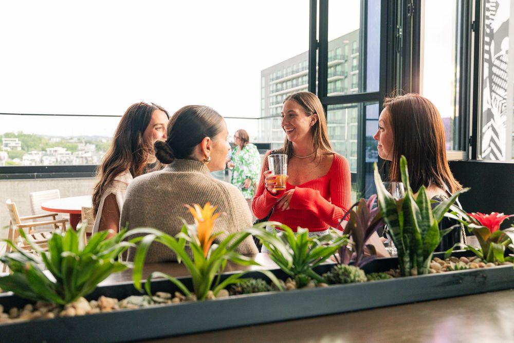 Four women socializing on a rooftop terrace surrounded by lush plants, enjoying drinks with a city view.