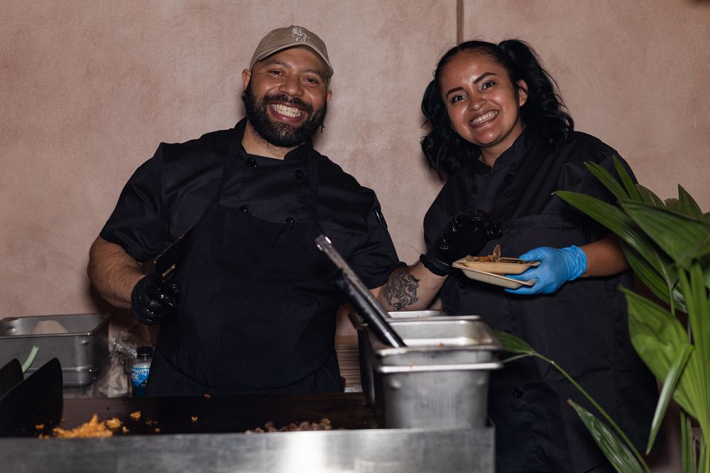 Two smiling chefs in black uniforms serve food at a catering event, one wearing blue gloves holding a plate.