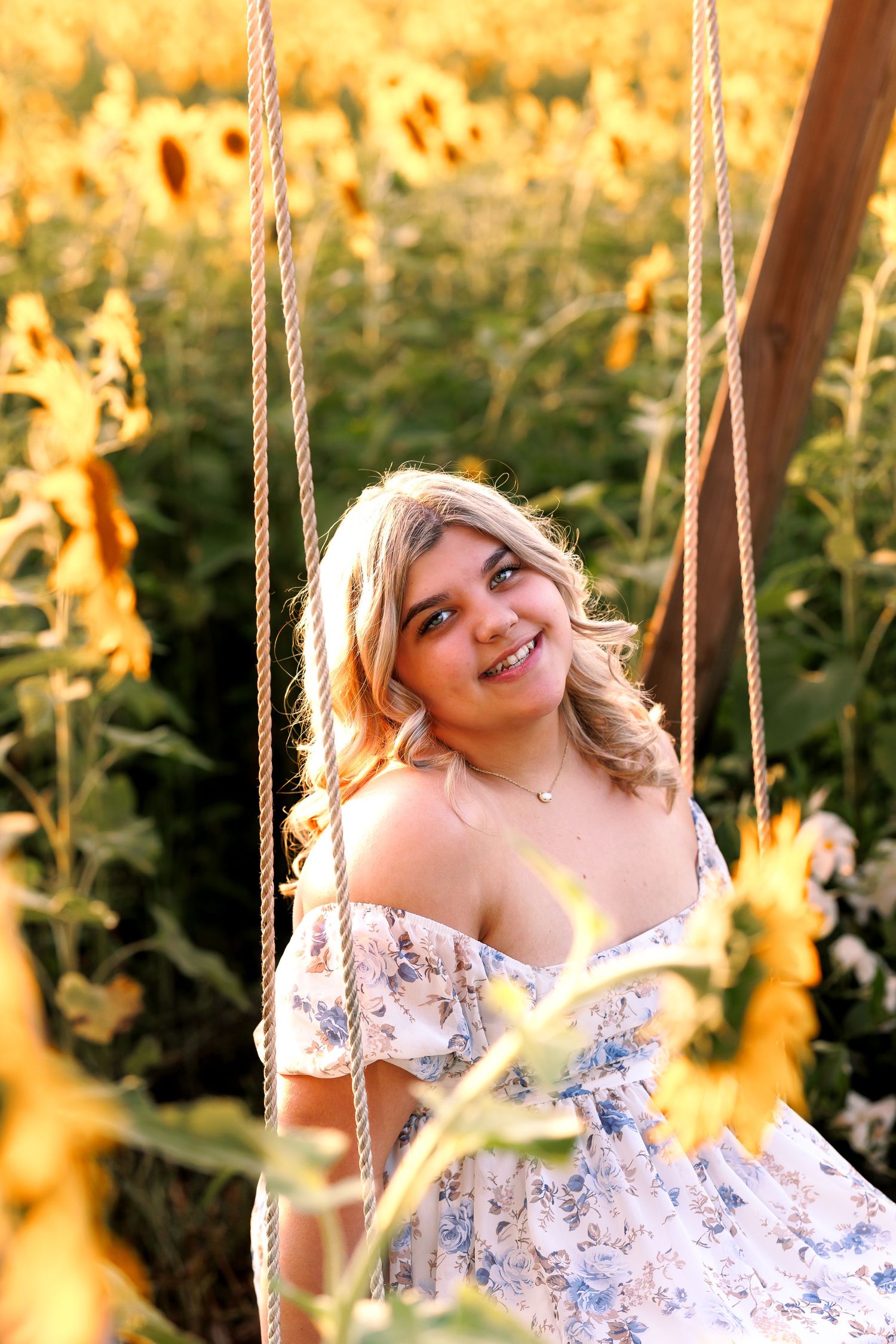 Person in floral dress sits on rope swing in sunlit garden during golden hour.