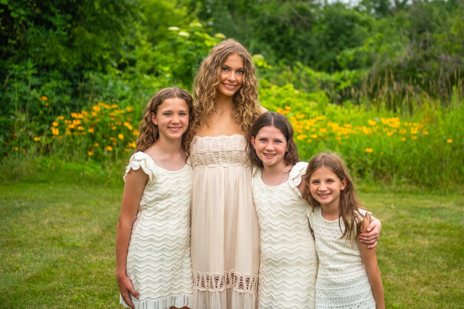 A group poses together in flowing cream-colored dresses against a summery backdrop of wildflowers and greenery.