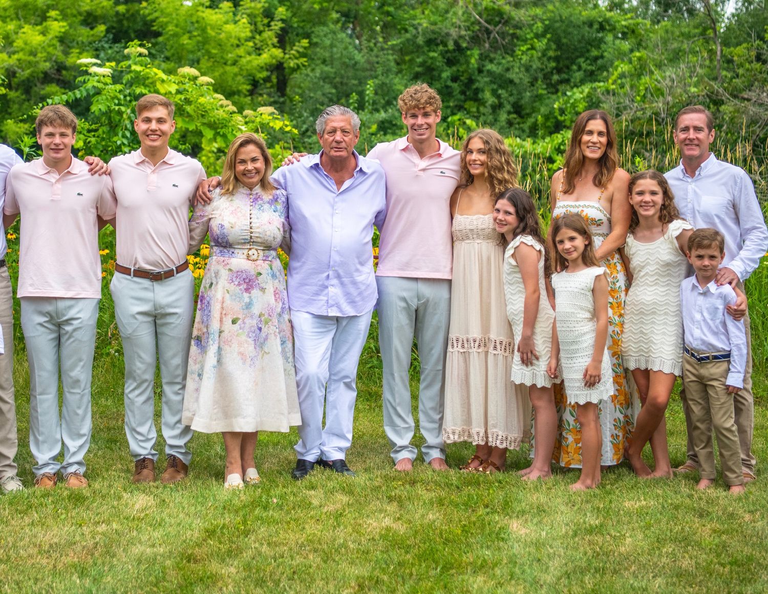 Extended family group dressed in white and beige casual clothing poses together outdoors in a lush green garden setting.